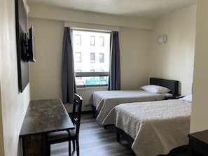 A hotel room with two beds, a desk, a chair, a window with curtains, and a view of buildings outside.