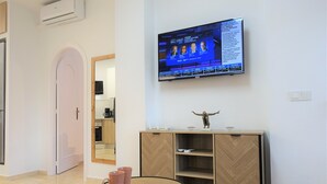 Ein modernes Hotelzimmer mit Fernseher, der Nachrichten zeigt, einem Holzschrank mit Fischgrätmuster und einem runden Tisch mit rosafarbenen Tassen.