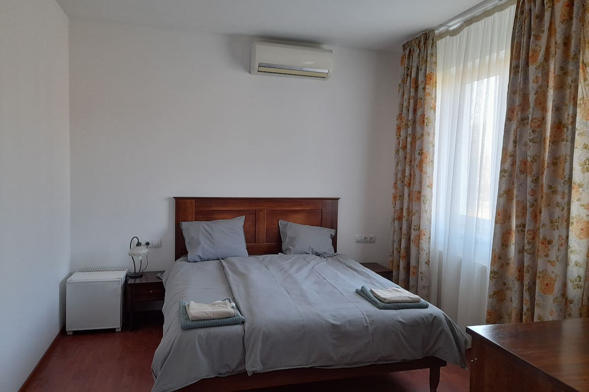 Un dormitorio con una cama de madera, una mesita de noche, una pequeña mesa y una ventana con cortinas florales.