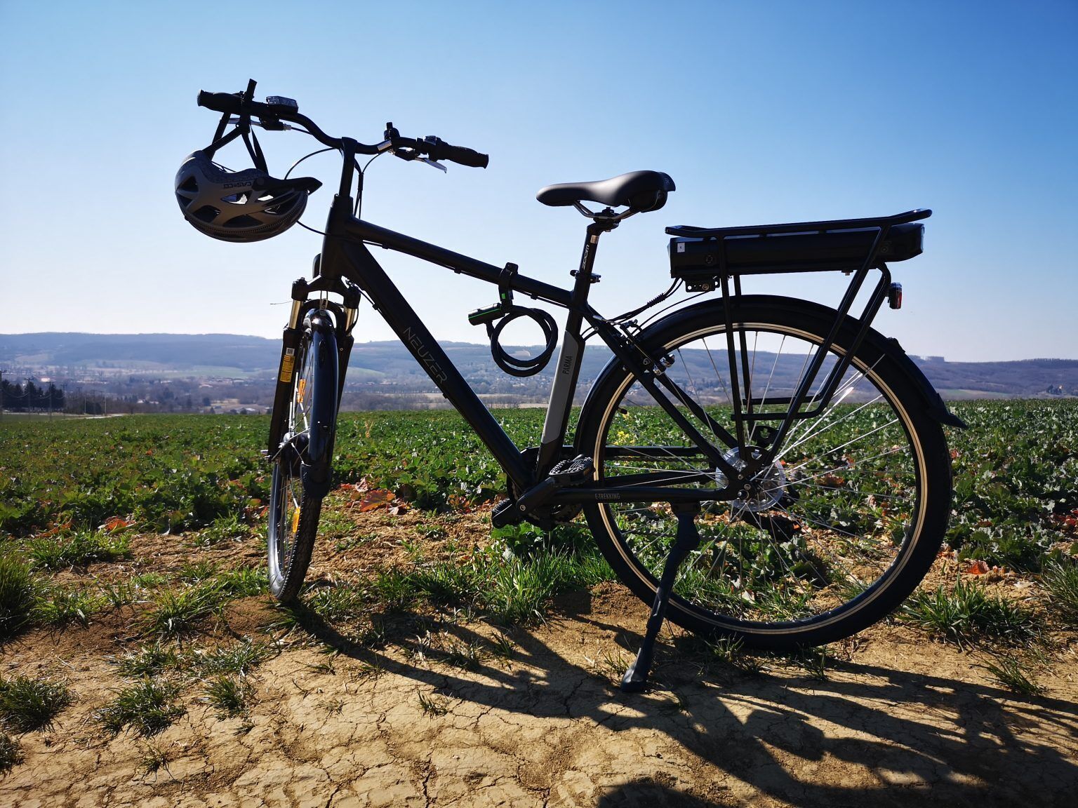 A bicycle with a front basket and a rear rack, parked on a dry, cracked ground with a field and distant hills in the background.
