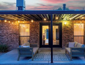 A covered outdoor seating area with brick walls, two sofas, and overhead string lights. - Rancho de Valle del Norte - An Irvie Home (Albuquerque)