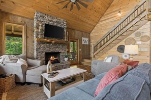 A cozy cabin interior with a stone fireplace, wooden walls, a flat-screen TV, and a staircase. - Mountain Views and Savings ~ Low Summer Rates! (Cherry Log)