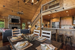 A wooden cabin interior with a dining area, kitchen, fireplace, and a staircase. - Mountain Views and Savings ~ Low Summer Rates! (Cherry Log)