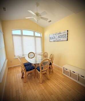 A dining area with a round glass table, four wicker chairs, a ceiling fan, and a wall sign.
