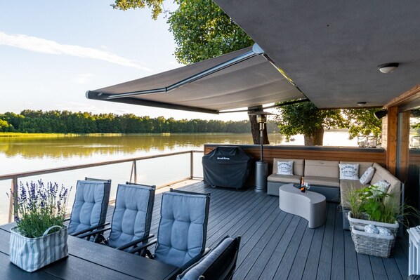 Una terraza con mobiliario exterior, un espacio cubierto y vistas a un lago.