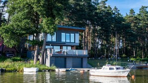 A modern houseboat with a large deck and a covered area, docked on a calm lake surrounded by trees.