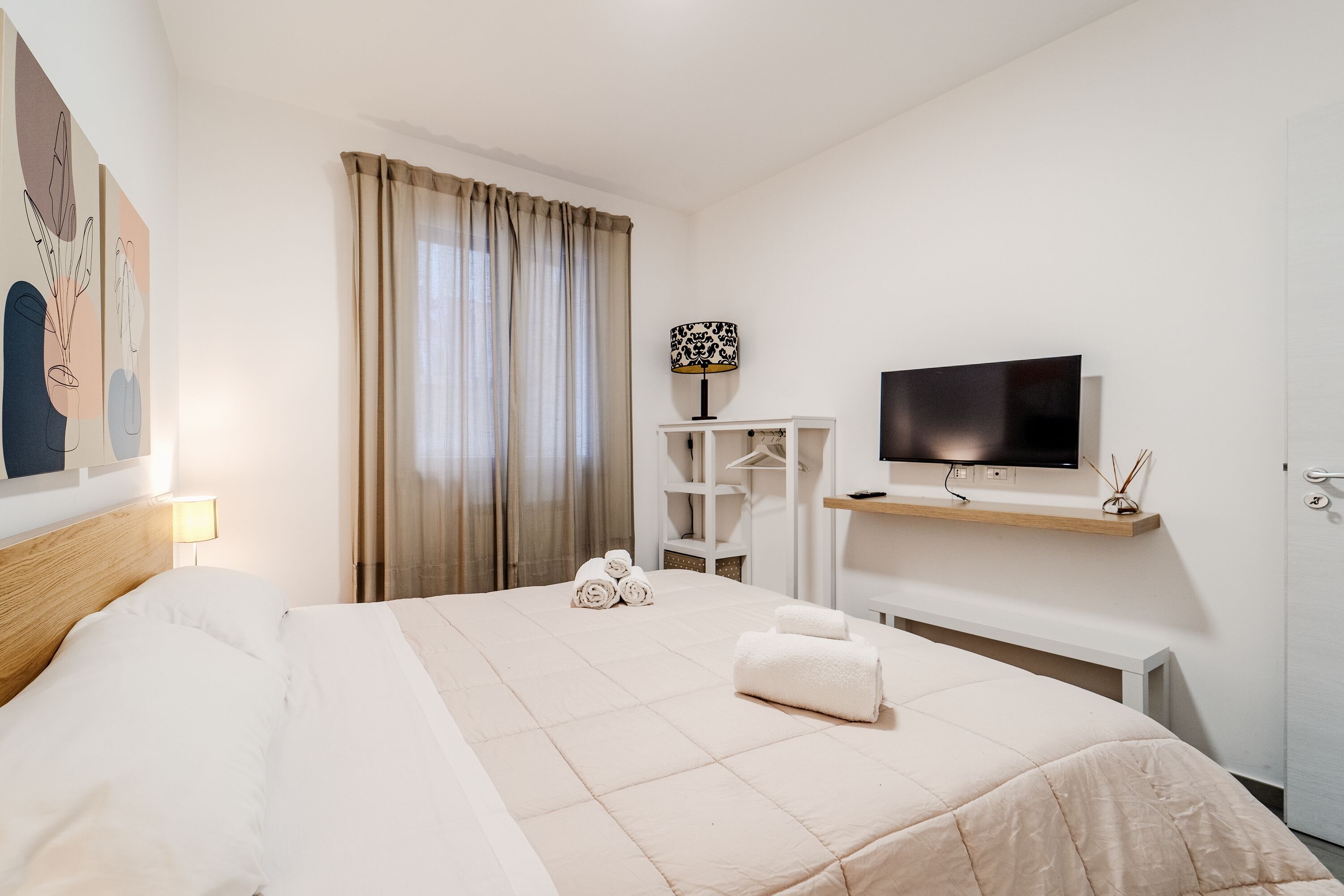 A hotel room with a bed, a TV, a desk, and a window with curtains.
