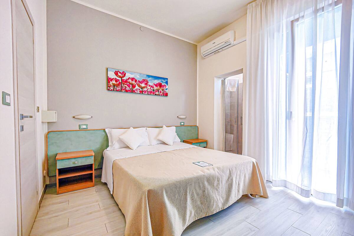 Een hotelkamer met een bed, een nachtkastje, een schilderij aan de muur, een raam met gordijnen en een deur.