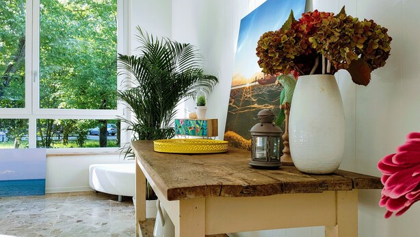 A room with a large window, a wooden table with a vase of flowers, a potted plant, and a decorative lantern.