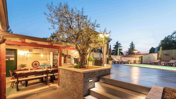 A well-lit outdoor area with a dining table and chairs, a stone planter with a tree, and a pool area with lounge chairs. - Holiday apartment with pool and air conditioning (Poreč)