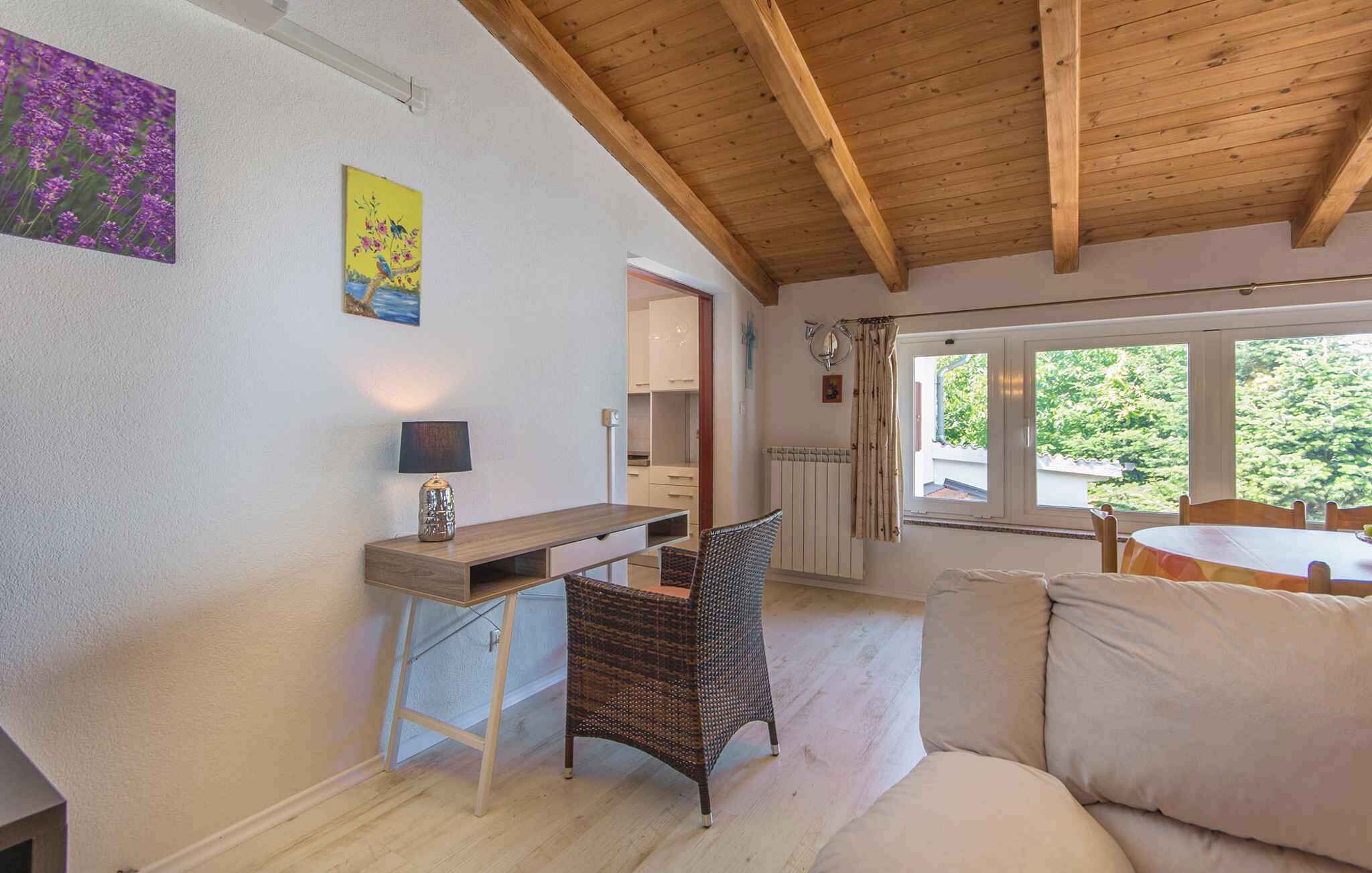 A cozy room with a wooden ceiling, a white sofa, a wicker chair, a small table with a lamp, and a window with a view of greenery.