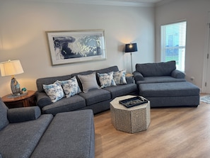 A living room with a sectional sofa, a round ottoman, a side table with a lamp, and a framed abstract painting on the wall.