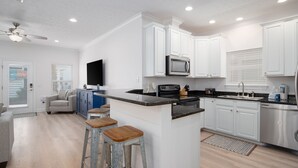 A modern kitchen with white cabinets, a black countertop, and a built-in microwave. There are wooden stools and a ceiling fan. - Laguna Beach PCB 5/3, STEPS TO THE BEACH-5 STAR RENTAL- CONVENIENT TO EVERYTHING (PANAMA CITY BEACH)