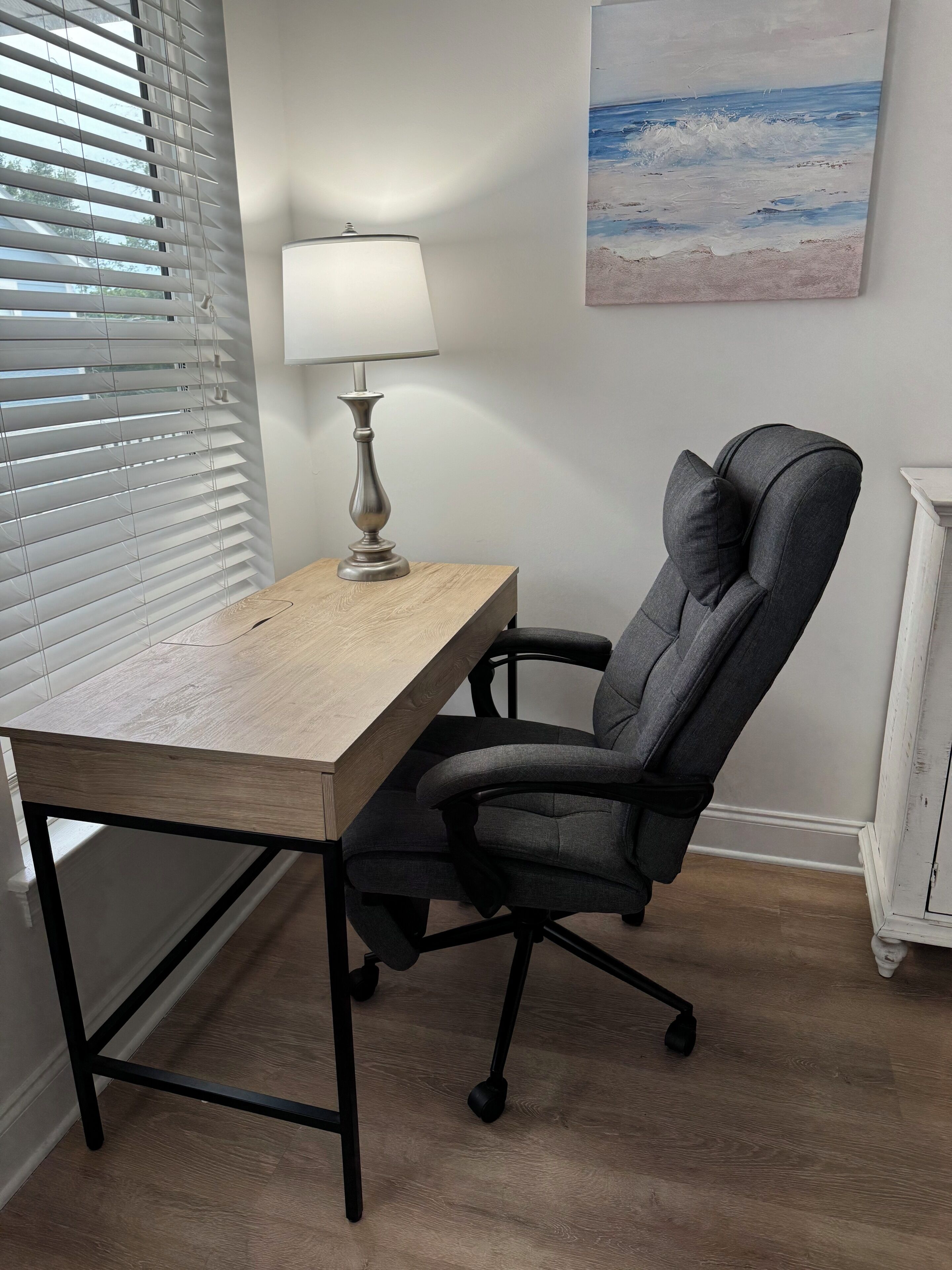 A room with a wooden desk, a modern office chair, a lamp, and a painting of a beach scene.