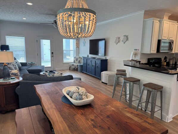 A modern living room with a wooden dining table, a large chandelier, and a kitchen area with white cabinets and a black countertop.