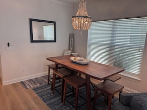 Un coin repas avec une table en bois, des bancs, un lustre, un miroir et une fen être avec des stores.