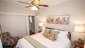 A bedroom with a bed, a ceiling fan, a floral artwork, a chair, and a nightstand with a lamp. - Near St. Joseph Hospital - Large Fenced Lot W/ W&D (Wichita)