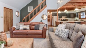 A cozy living room with a brown leather sofa, a grey couch, and a wooden coffee table. A wooden staircase leads to an upper level with a kitchen area visible. - Pocono Lake Chalet w/Hot Tub, Pool Table, & Fire Pit. Pet friendly! (Pocono Lake)