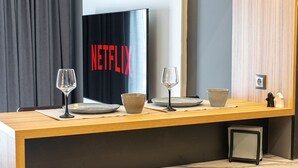 Una moderna camera d'albergo con un tavolo da pranzo in legno apparecchiato per due persone, con un bicchiere da vino in vetro, una ciotola in ceramica e un piatto. Si vede a parete un'ampia cornice con il logo di Netflix.