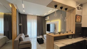 Un soggiorno moderno con un divano, un tavolino da caffè e una televisione. È presente una parete divisoria in legno con un piano in marmo e un piccolo tavolo con bicchieri da vino.