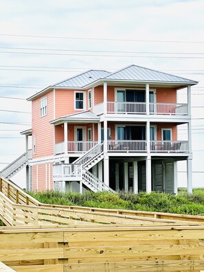 A multi-story beachfront house with a balcony, wooden deck, and a staircase leading to the entrance.