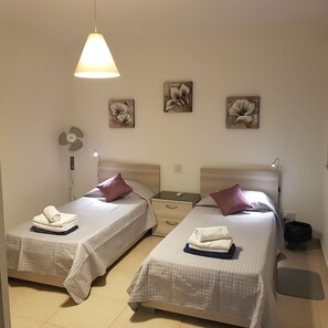 Two single beds with white bedding and pillows, a bedside table, a wall-mounted fan, and a pendant light.