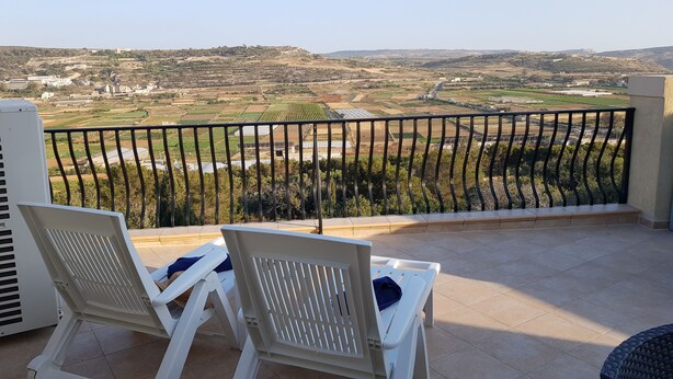 Ein Balkon mit zwei weißen Stühlen und Blick auf terrassenförmig angelegte Felder sowie entfernte Gebäude.