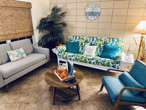 Una sala de estar con un sofá gris, un sillón azul, una mesa de centro redonda de madera y una palmera decorativa.