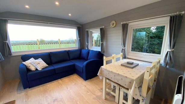 Ein Zimmer mit einem blauen Modulsofa, einem kleinen Tisch mit Stühlen und großen Fenstern mit Blick auf Grünflächen.