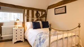 A bedroom with a white bed, a wooden headboard, a nightstand with a lamp, and a window with a view of a brick building. - Miller Cottage - A gorgeous historic cottage in picturesque Saffron Walden (Saffron Walden)