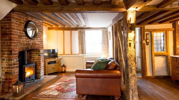 A cozy living room with a brick fireplace, wooden beams, a leather sofa, and a rustic stone pillar. - Miller Cottage - A gorgeous historic cottage in picturesque Saffron Walden (Saffron Walden)