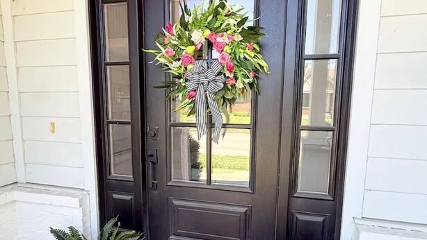 Una puerta de madera oscura con un adorno de lazo, un felpudo que dice 'hello' y dos macetas con plantas.