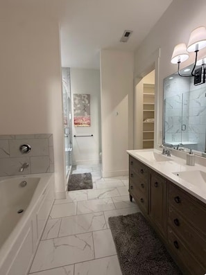 A modern bathroom with a bathtub, vanity with a large mirror and lights, marble tiles, and a hallway leading to another room. - Downtown Bourbon Black Swan House (Bardstown)