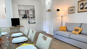 A modern living room with a grey sofa, a dining table set with plates and glasses, a bookshelf, and a flat-screen TV. - HAPPY DAYS large ground floor apartment with garden and parking. (Intra)