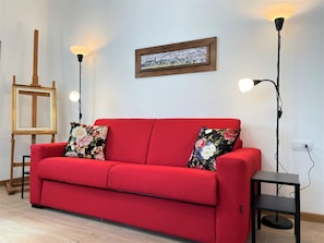 A red sofa with floral cushions, a framed picture on the wall, and two floor lamps.