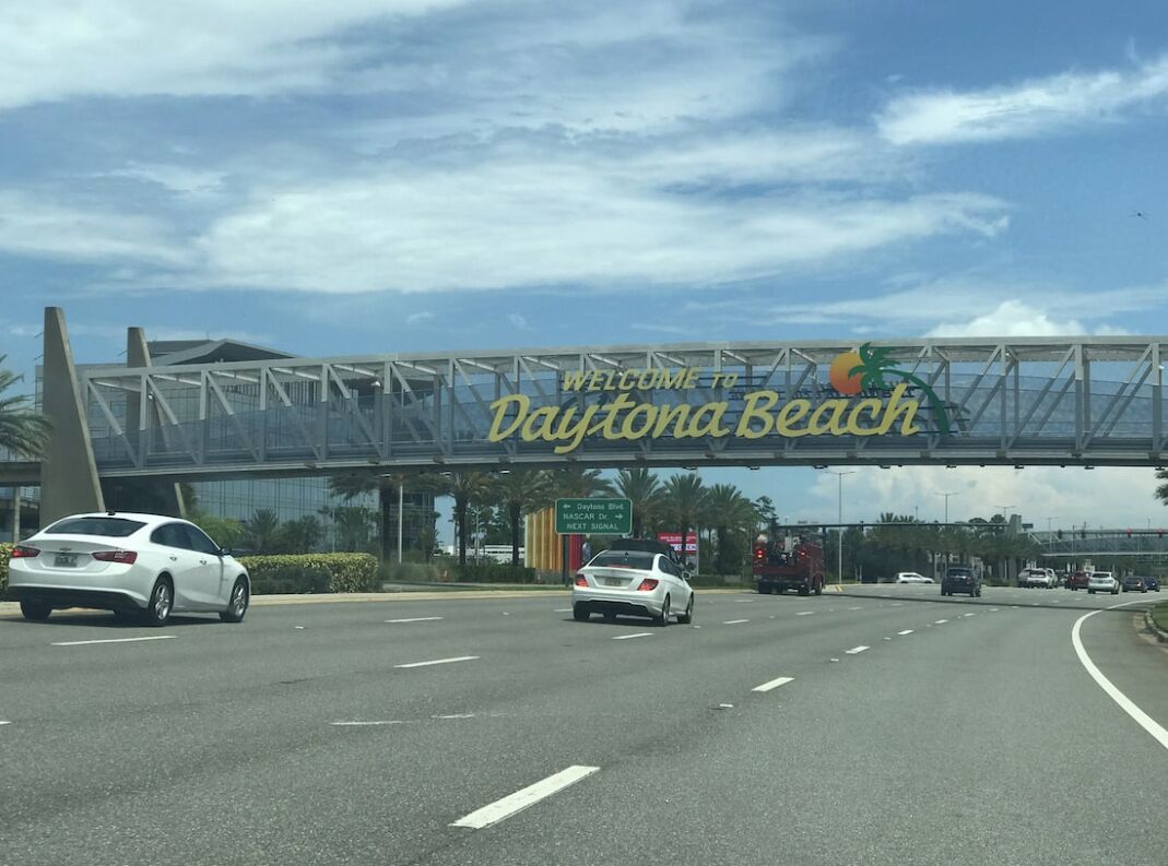 A road sign welcoming visitors to Daytona Beach with a clear sky and multiple vehicles on the road.