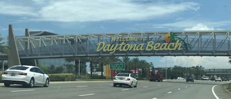 A road sign welcoming visitors to Daytona Beach with a clear sky and multiple vehicles on the road.