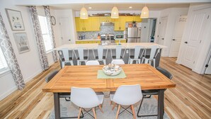 A dining area with a wooden table and white chairs, a kitchen in the background, and a clock on the wall. - 2nd Row Beach House just steps to the sand!  EV  charger! (Surfside Beach)