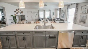 A modern kitchen with grey cabinetry, a large island with a sink, pendant lights, and a dining area with chairs. - 2nd Row Beach House just steps to the sand!  EV  charger! (Surfside Beach)