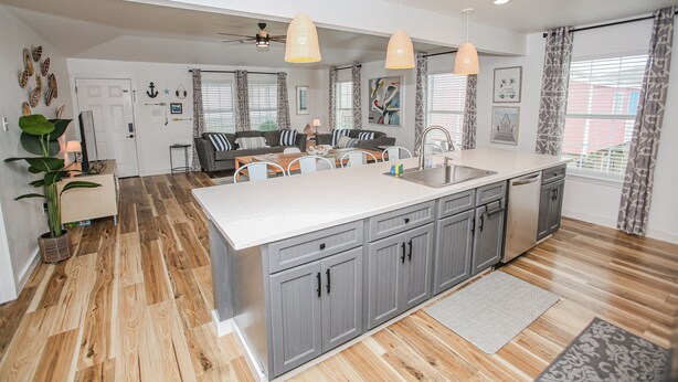 A modern kitchen with a large island, grey cabinets, and a white countertop. There is a dining area with a table and chairs, and a kitchen island with a sink and dishwasher. - 2nd Row Beach House just steps to the sand!  EV  charger! (Surfside Beach)