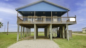 A blue wooden structure with a balcony, supported by wooden stilts, overlooking a grassy area. - 2nd Row Beach House just steps to the sand!  EV  charger! (Surfside Beach)