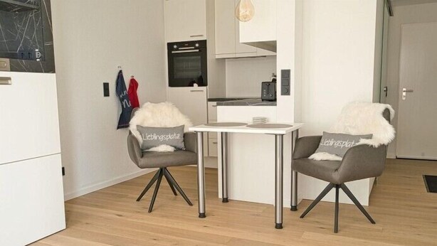 Een moderne keuken met een inbouwoven en een zithoek met twee grijze stoelen en een kleine tafel.