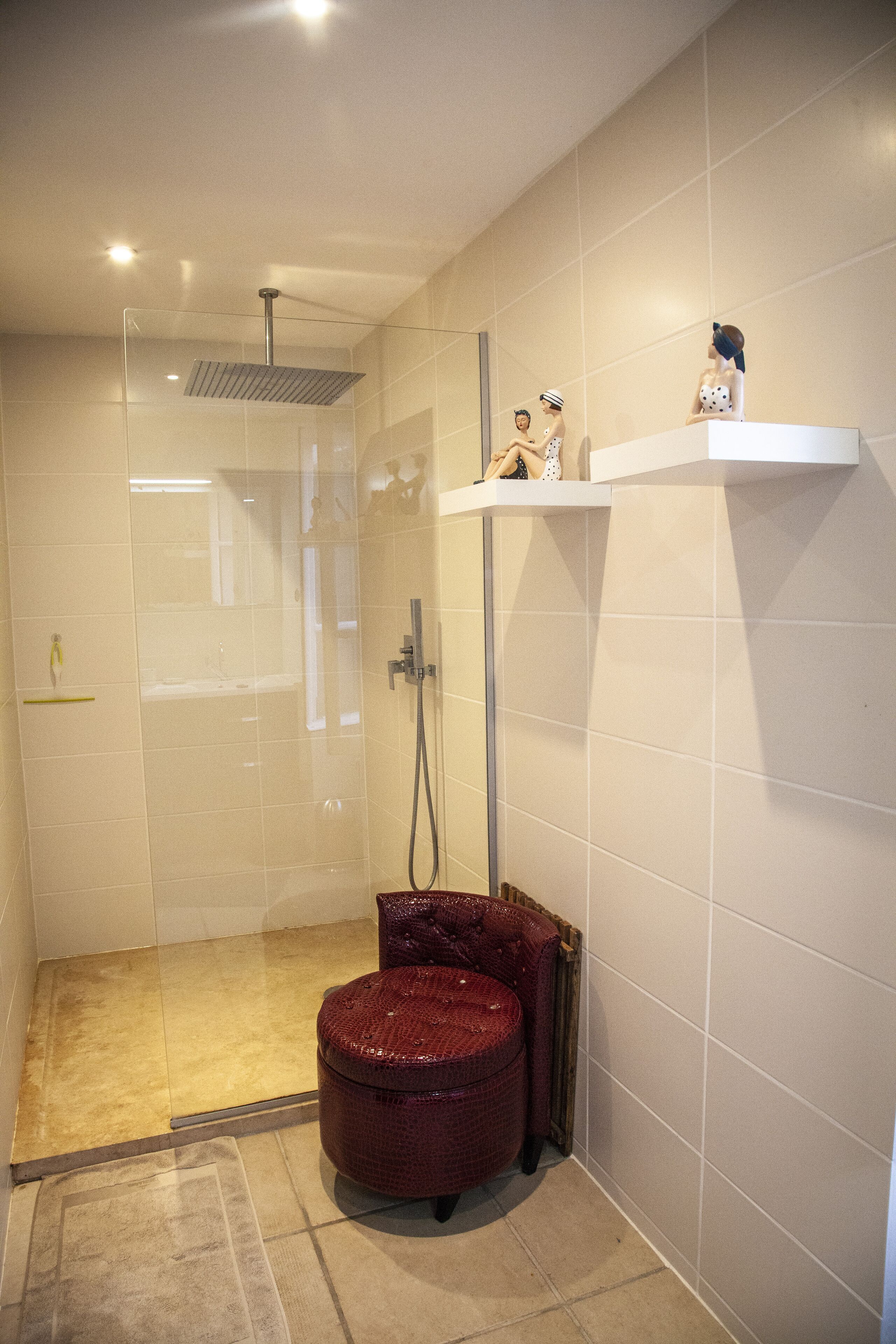A modern bathroom with a glass-enclosed shower, a red stool, and a shelf with decorative items.
