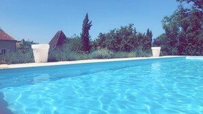 Une piscine d’un bleu éclatant, bordée de deux jardinières blanches, entourée de verdure et d’un bâtiment dont le toit est recouvert de tuiles.