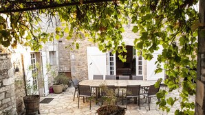 Une terrasse extérieure avec un sol en pierre, une table en bois et des chaises. On y trouve des plantes en pot et, au fond, un mur en pierre.