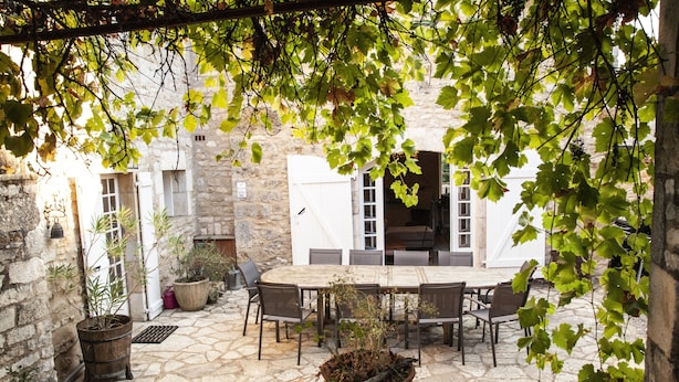 An outdoor patio with a stone floor, a wooden table, and chairs. There are potted plants and a stone wall in the background.