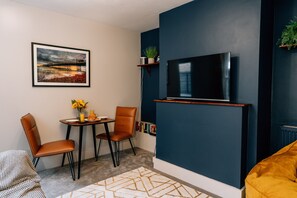 Uma sala de estar moderna com televisão de tela plana presa a uma parede azul escuro, uma área de jantar com duas cadeiras e uma mesa pequena, e um sofá.
