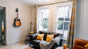 Un salon avec un canapé en cuir foncé, une guitare, une table basse sur laquelle se trouve un bol de citrons, et un coin repas avec une assiette de croissants et un verre de jus d’orange.