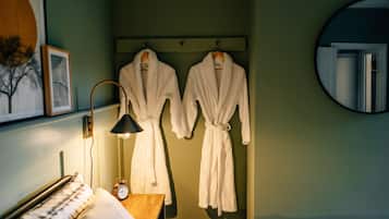 A hotel room with a bed, a bedside table with a lamp, a clock, and two white bathrobes hanging on a rack.