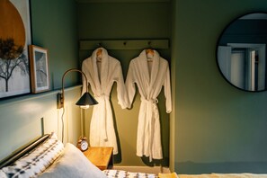 A hotel room with a bed, a bedside table with a lamp, a clock, and two white bathrobes hanging on a rack. - Sanctuary-by-Sea  2 bed Apartment (Worthing)